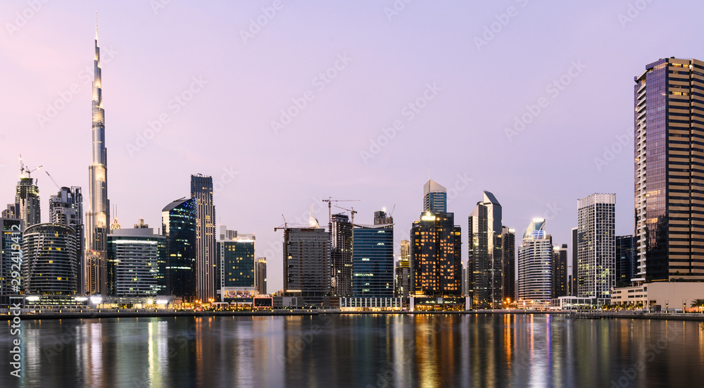 Stunning view of the illuminated Dubai skyline during sunset with tall skyscrapers and a water canal flowing in the foreground. Dubai, United Arab Emirates.
