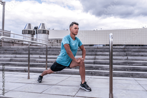 Male athlete guy, in summer in city, doing warm-ups of knees of joints and legs, ankle-boot, before morning jogging, fitness workout training, background concrete steps of cloud.