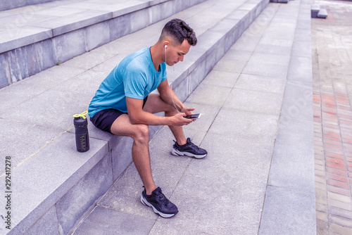 male athlete, tanned man, summer city, resting his hands with mobile phone, listening music headphones, bottle protein water, fitness workout morning joggings. Free space for motivation text.