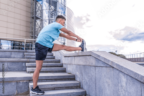 Male athlete, tanned man, summer in city, view from side, stretching muscles of legs knees ankle, warming up before jogging, fitness training workout. Free space for motivation text.