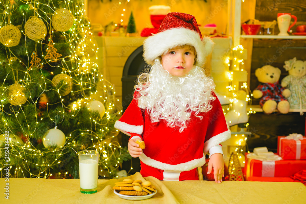 Cookies for kids Santa Claus. Portrait of little Santa child holding chocolate cookie and glass of milk. Santa kids picking cookie. Little Santa picking cookie and glass of milk at home.
