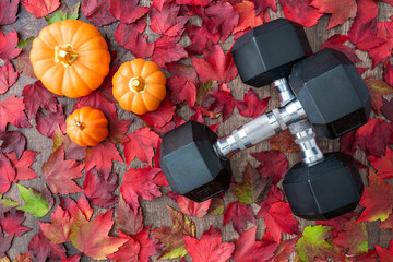 Pair of crossed dumbbells on a rustic wood background covered in fall color of red, green, yellow, and orange maple leaves, with ceramic pumpkins
