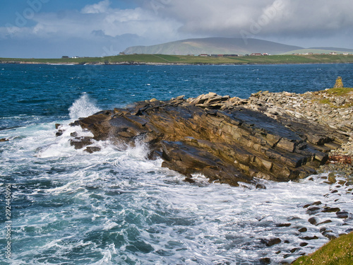 Wave action on inclined sandstone rocks on the south Shetland coast near Sumburgh Head, showing rock debris swept by waves on the right. The hill of Fitful Head is in the background.