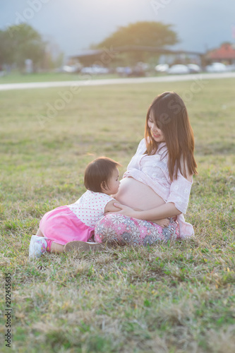 baby girl hugging asian beautiful pregnant woman on grass field in evening light with sunset and mountain background, Good health and relax concept, family relationship.