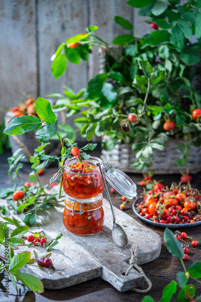 Rose hip jam in jar pots on wooden board. Ripe red berries with leaves in plate and basket. Autumn harvesting season, fresh vitamins on gray background. Healthy nutrition, homemade marmalade concept