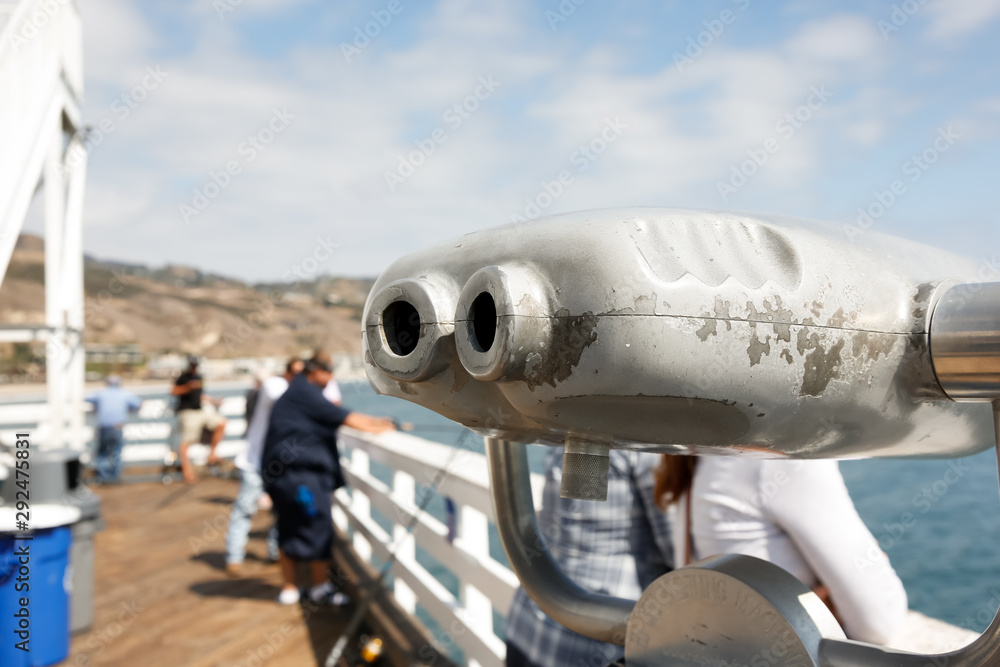 A closeup view of pier binoculars, featuring people in the background fishing and enjoying the view. A closeup view of pier binoculars, featuring people in the background fishing and enjoying the view.