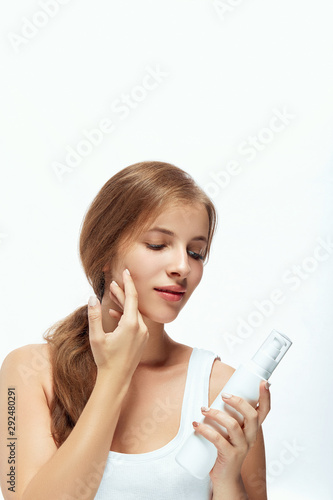 Beauty Woman Face Skin Care. Portrait Of Attractive Young Female Applying Cream And Holding Bottle. Closeup Of Smiling Girl With Natural Makeup And Fresh Skin. Beauty Cosmetics.