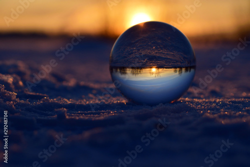 Glass sphere with reflection of sunset standing on snow surface. Purple colors. Winter nature background