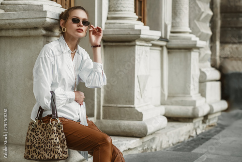 Outdoor autumn fashion portrait of elegant, luxury woman wearing sunglasses, trendy white shirt, leather trousers, holding animal, leopard print handbag, posing in street of European city. Copy space
