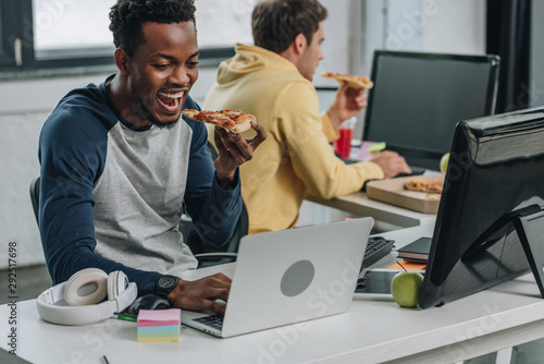 two multicultural programmers eating pizza while sitting near computers in office