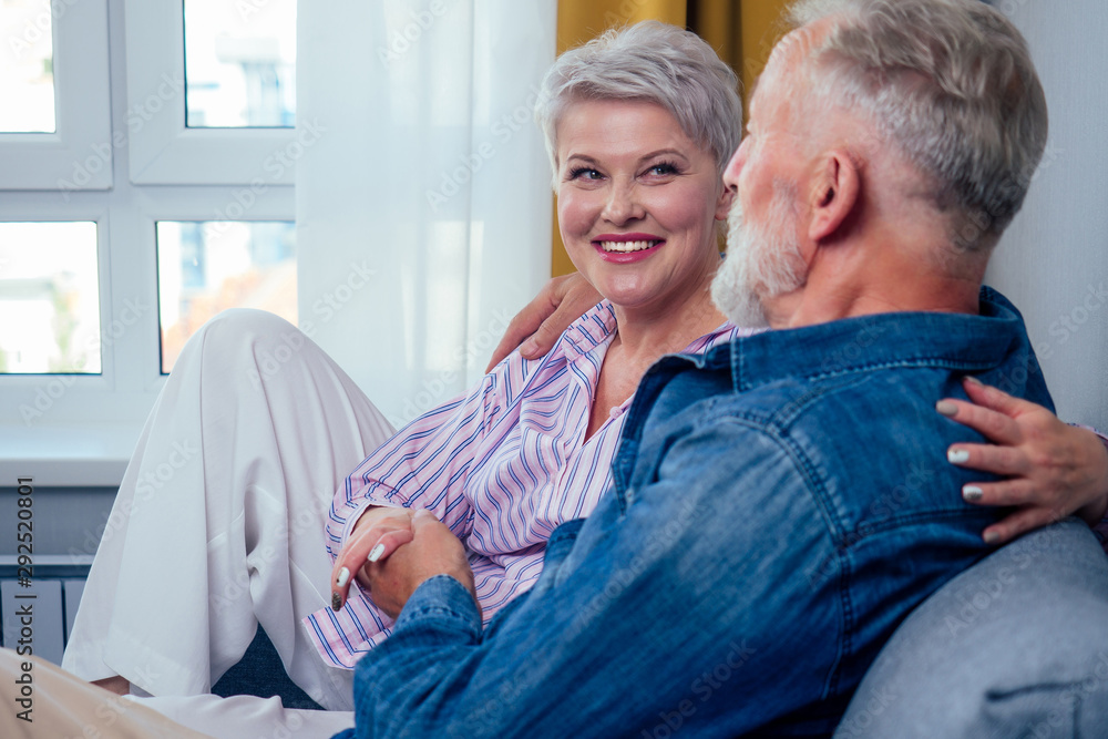 Mature good-looking well dressed couple sitting in sofa in day light from big window and yellow curtains in living room,touching hand Mature good-looking well dressed couple sitting in sofa in day light from big window and yellow curtains in living room,touching hand