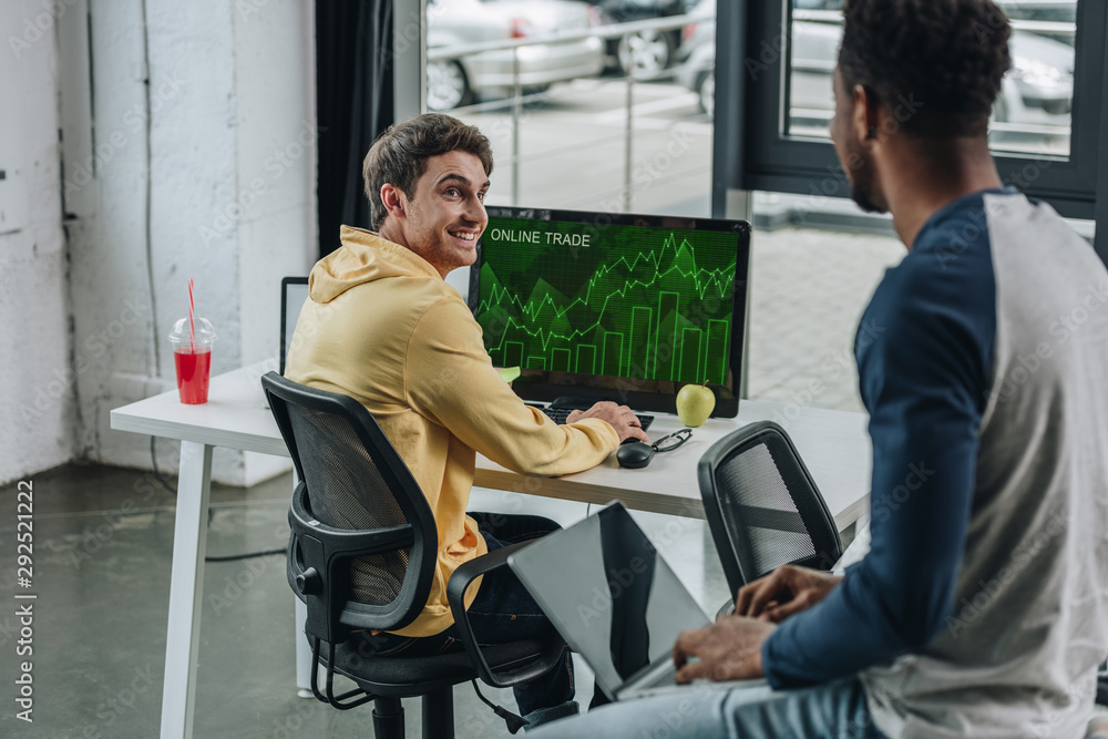 cheerful programmer sitting at computer monitor with online trade on screen and looking at african american colleague cheerful programmer sitting at computer monitor with online trade on screen and looking at african american colleague