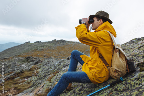 stylish hikers in raincoat, green hat with backpack holding binoculars sitting on top of the mountain and looks into the distance.close up side view photo. copy space