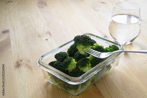 Steamed boiled broccoli on a fork in a glass container and pure water on a wooden background with copy space. Proper nutrition, super food and fitness