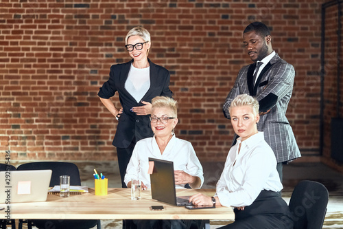 Three caucasian adult blond woman wear white shirt and one of them white shirt and black jacket, and one african man wear grey checkered jacker move, sit in office room photo