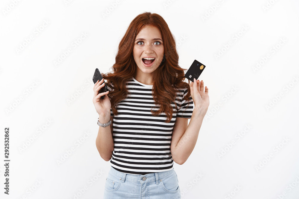 Excited attractive redhead girl raising smartphone and credit card, smiling fascinated looking upbeat, enjoy shopping online from home, input banking number, purchase outfit for prom