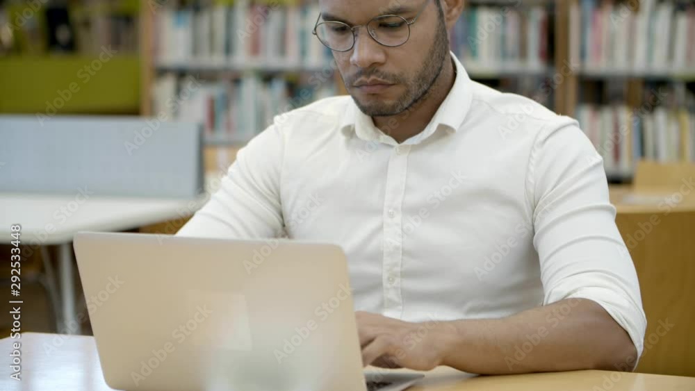 Concentrated student in eyeglasses typing on laptop. Front view of handsome young man working with laptop at library. Education concept Concentrated student in eyeglasses typing on laptop. Front view of handsome young man working with laptop at library. Education concept