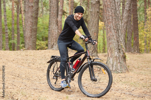 Pictiure of biker riding through forest  wearing black cycling clothes  cap and sneakers  having happy facial expression  sporty male spending leisure time in open air  riding around trrees.