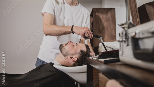 Young man with trendy haircut at barber shop. Barber washes customer head.
