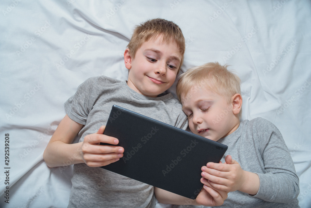 Two boys lying in bed and watching something on a tablet. Gadget Leisure Two boys lying in bed and watching something on a tablet. Gadget Leisure