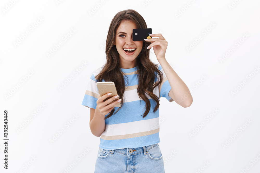 Amused cheerful european moder girl like ordering products online, booking flight smartphone app, hold mobile phone and credit card near eye, smiling joyfully, buy in internet store, white background Amused cheerful european moder girl like ordering products online, booking flight smartphone app, hold mobile phone and credit card near eye, smiling joyfully, buy in internet store, white background