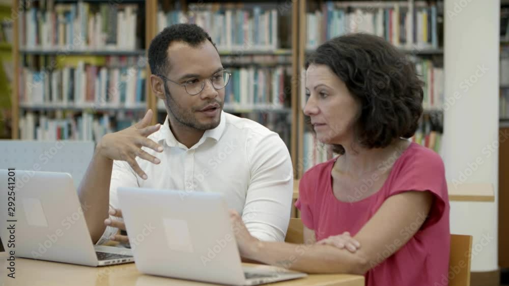 Slow motion shot of two people talking at library. Focused students sitting at table with laptops and discussing new project. Education concept Slow motion shot of two people talking at library. Focused students sitting at table with laptops and discussing new project. Education concept