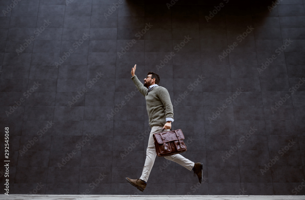 Full length of handsome bearded caucasian fashion designer holding leather bag and stopping taxi.