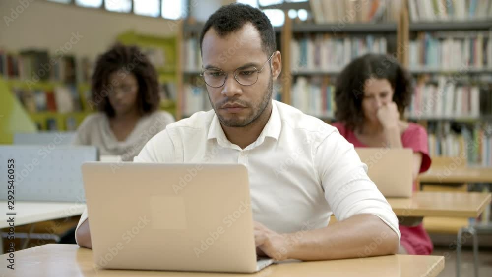 Front view of African American man using laptop at library. Thoughtful African American student in eyeglasses sitting at table and typing on laptop. Education concept Front view of African American man using laptop at library. Thoughtful African American student in eyeglasses sitting at table and typing on laptop. Education concept