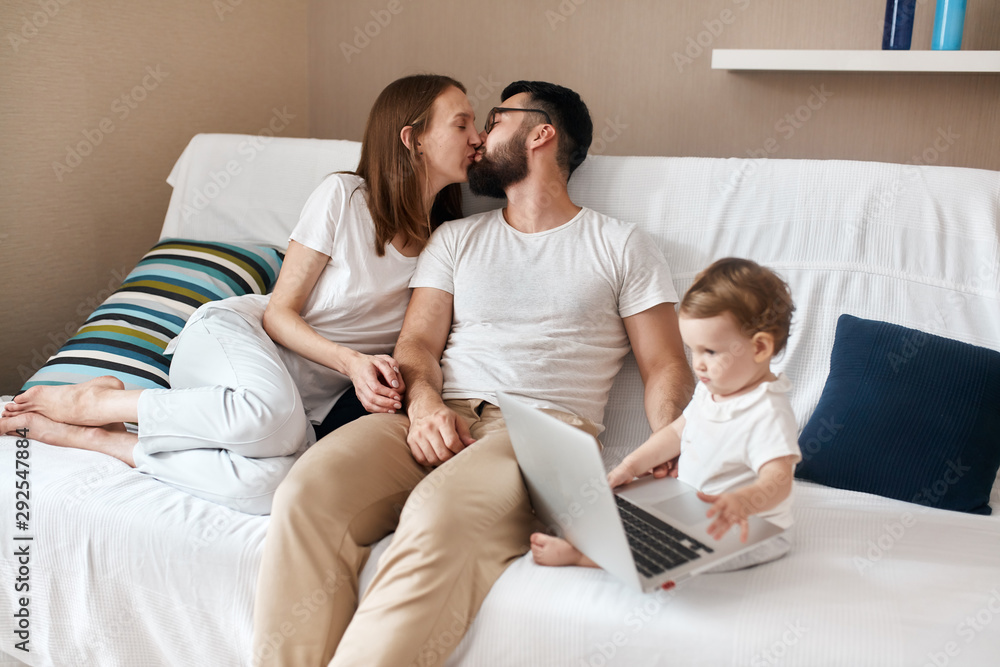 loving couple kissing while their baby watchcing cartoon on the laptop, love, warm, tender feeling and emotion loving couple kissing while their baby watchcing cartoon on the laptop, love, warm, tender feeling and emotion