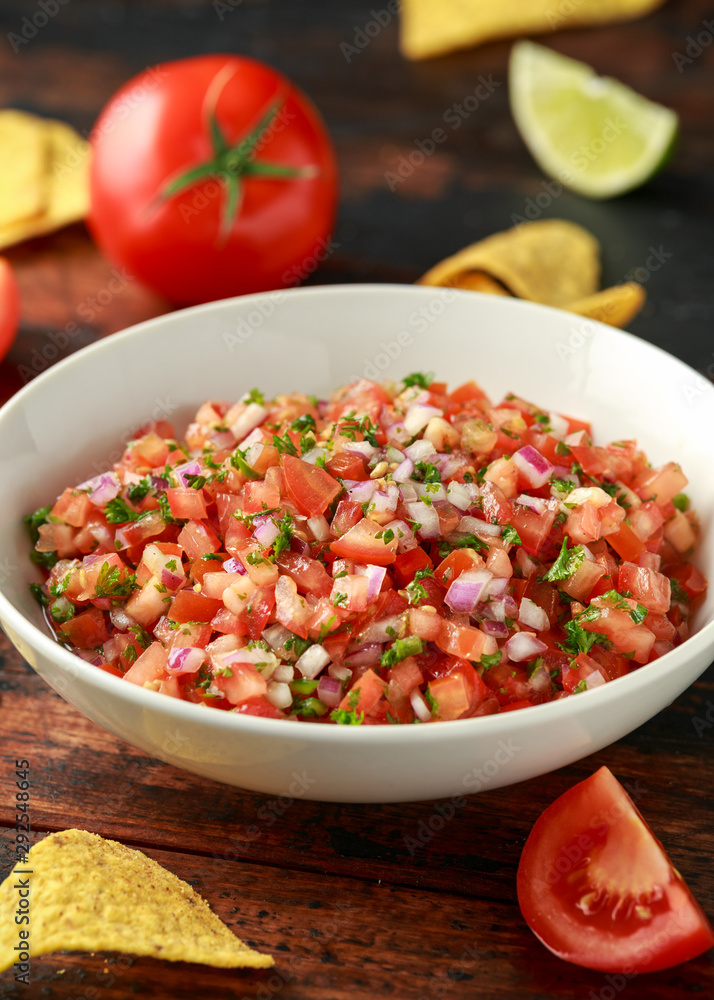 Mexican Tomato Salsa in white bowl with lime, red onion, jalapeno pepper, parsley and tortilla chips on wooden table Mexican Tomato Salsa in white bowl with lime, red onion, jalapeno pepper, parsley and tortilla chips on wooden table