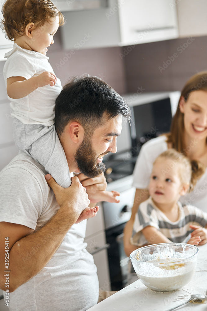 happy father and baby who is sitting on his shoulder controlling their mommy's work in the kitchen, close up side view cropped photo happy father and baby who is sitting on his shoulder controlling their mommy's work in the kitchen, close up side view cropped photo