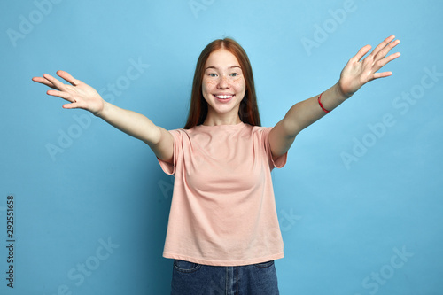 Beauitul positive girl wearing pink t-shirt over isolated blue background looking at the camera smiling with open arms for hug. Cheerful expression. close up portrait, studio shot photo