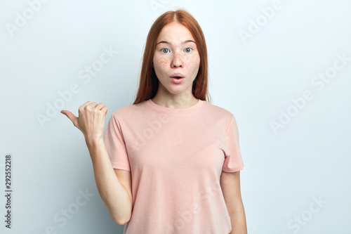shocked scared beautiful freckled young woman with foxy long straight indicates with thumb acopy space, shows something strange aside, isolated on white.look here. body language photo