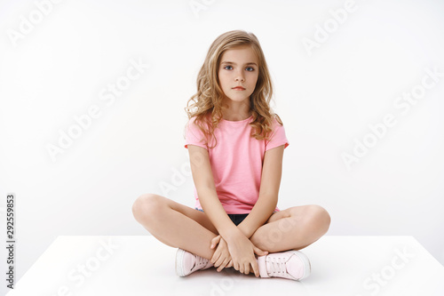 Canvastavla Serious indifferent cute blond little girl sitting on floor, legs crossed, look