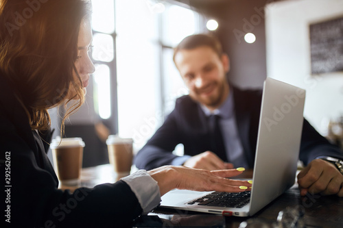 young woman's hands on laptop. serious attractive brown-haired woman concentretaed on working with laptop, while her colleague watching her, blurred background © the faces