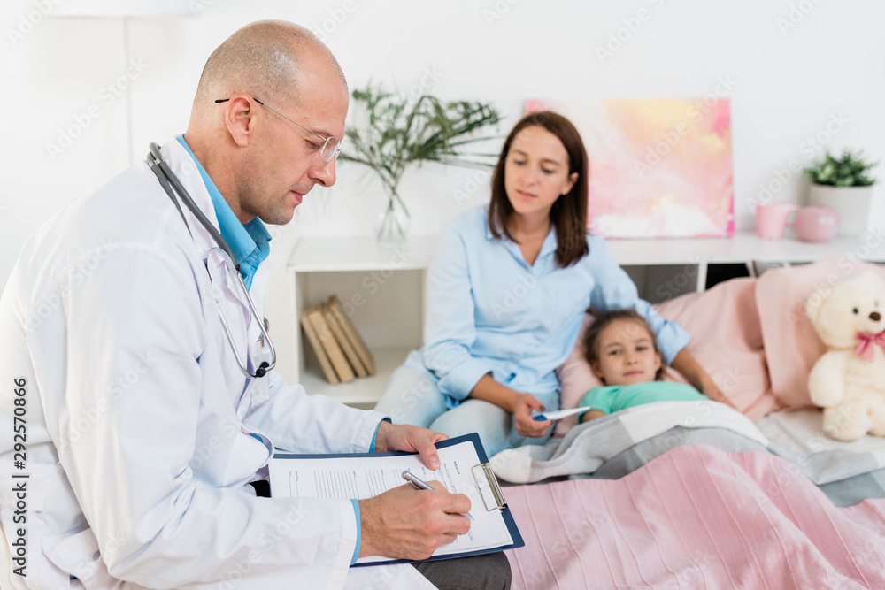 Serious doctor making prescriptions to sick little girl while sitting by her bed Serious doctor making prescriptions to sick little girl while sitting by her bed