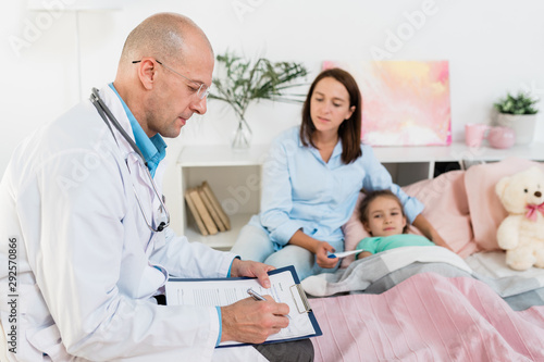 Serious doctor making prescriptions to sick little girl while sitting by her bed