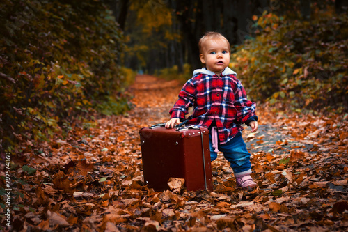 Cheerful toddler hipster toddler in a red checkered shirt holds a suitcase, looks at the camera and smiles. Autumn park and a road with fallen leaves. Full-length Artistic Portrait © Natal.is