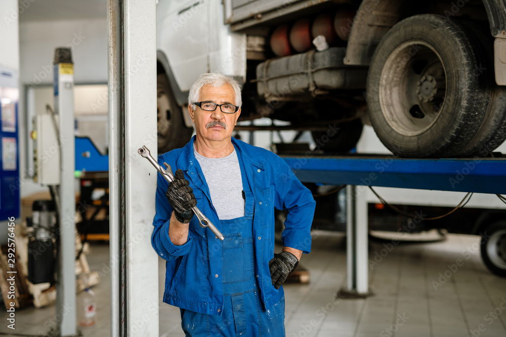 Serious grey-haired man in workwear standing in workshop or hangar Serious grey-haired man in workwear standing in workshop or hangar