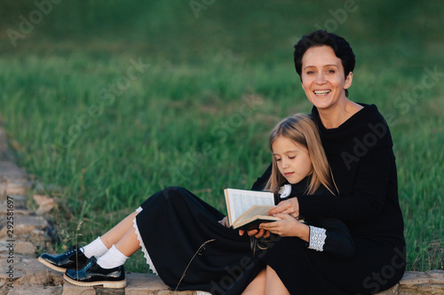 Mother and daughter are sitting on a stone bench and reading a book. Woman with a child in black dresses.