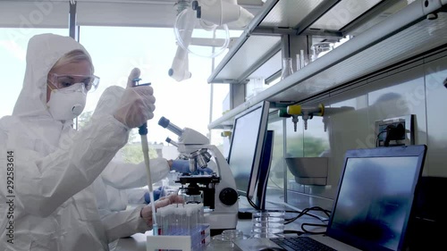 Scientists in protection suits and masks working in research lab using laboratory equipment: microscopes, test tubes. Biological hazard, pharmaceutical discovery, bacteriology and virology. photo