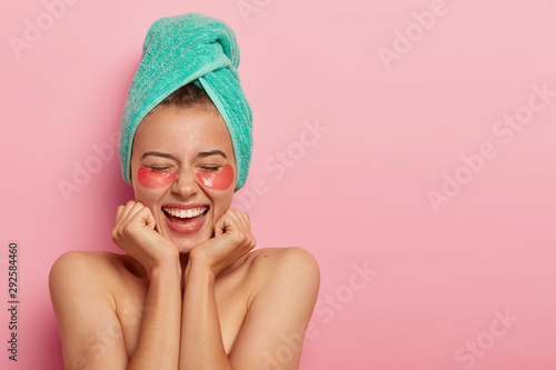 Photo of overjoyed European woman touches chin with both hands, shows bare shoulders, has smooth healthy skin, removes wrinkles under eyes with cosmetic patches, isolated over rosy studio wall