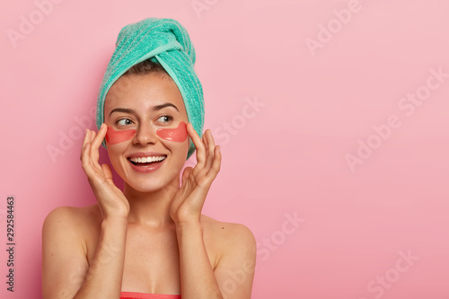 Image of cheerful woman touches gently face, smiles pleasantly, applies hydrogel patches under eyes, stands naked, has healthy skin, isolated over pink background, blank space for promotional content
