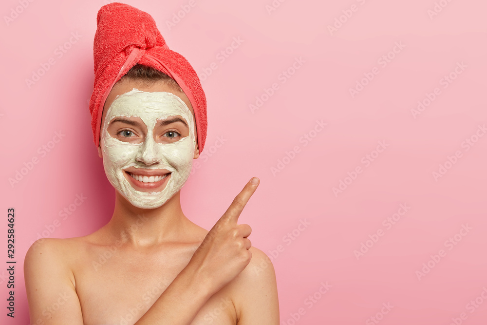 Studio shot of cheerful young woman with gentle smile, points index finger on copy space, advertises beauty item, stands shirtless, wears clay mask and towel poses against pink background. Cosmetology Studio shot of cheerful young woman with gentle smile, points index finger on copy space, advertises beauty item, stands shirtless, wears clay mask and towel poses against pink background. Cosmetology