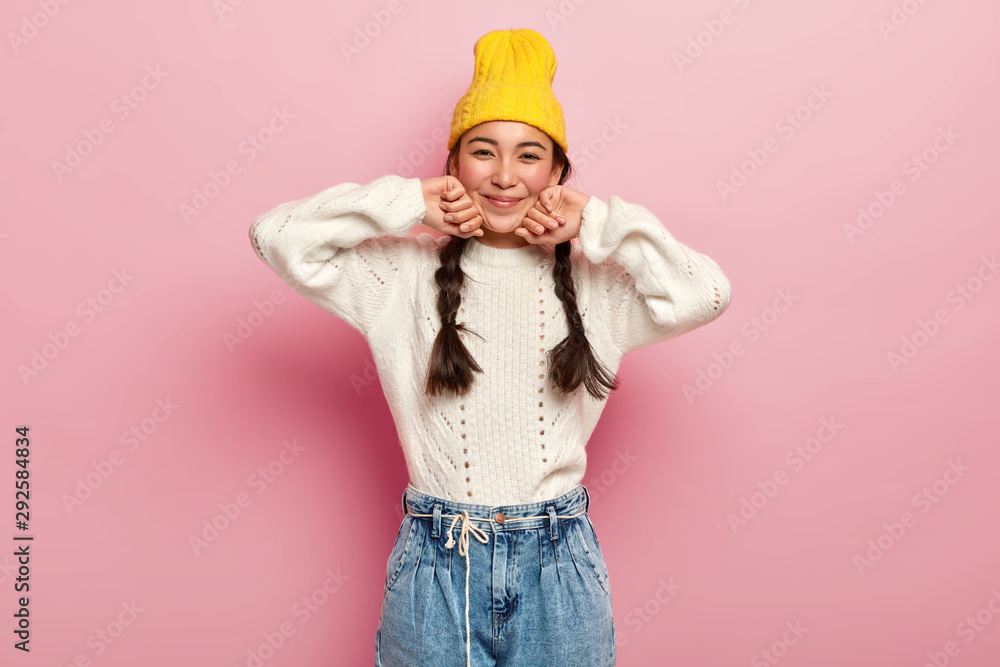 Pretty mixed race female dressed in fashionable yellow hat, white sweater and jeans, has enthusiastic and charming look in camera, glad expression, isolated over pink background, has two combed plaits - obrazy, fototapety, plakaty 