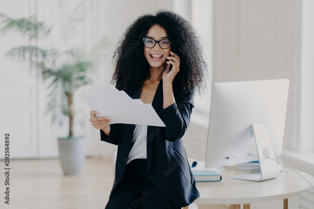 Obraz premium Intelligent woman with Afro hairstyle, holds paper documents, checks information, calls business partner via cell phone, analyzes future strategy, speaks with banking manager, dressed in black suit