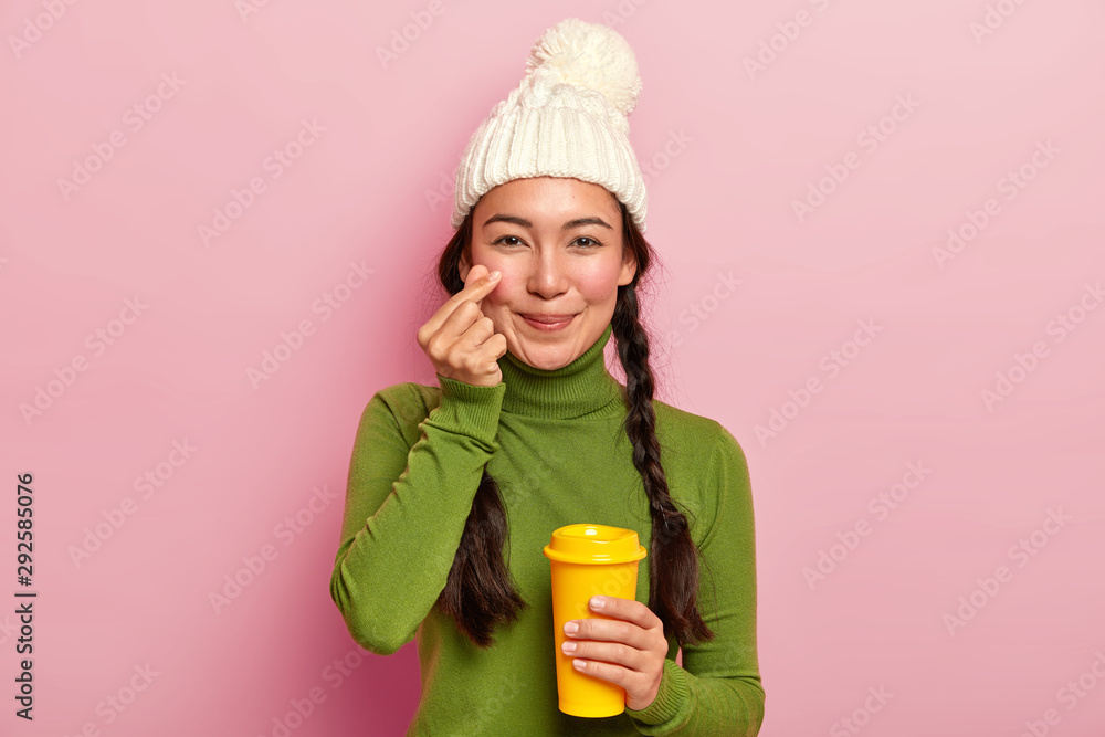 Photo of attractive young Asian girl makes korean like sign, shapes little heart near cheek, expresses love to boyfriend, wears warm hat and casual jumper, drinks takeaway coffee, poses indoor Photo of attractive young Asian girl makes korean like sign, shapes little heart near cheek, expresses love to boyfriend, wears warm hat and casual jumper, drinks takeaway coffee, poses indoor