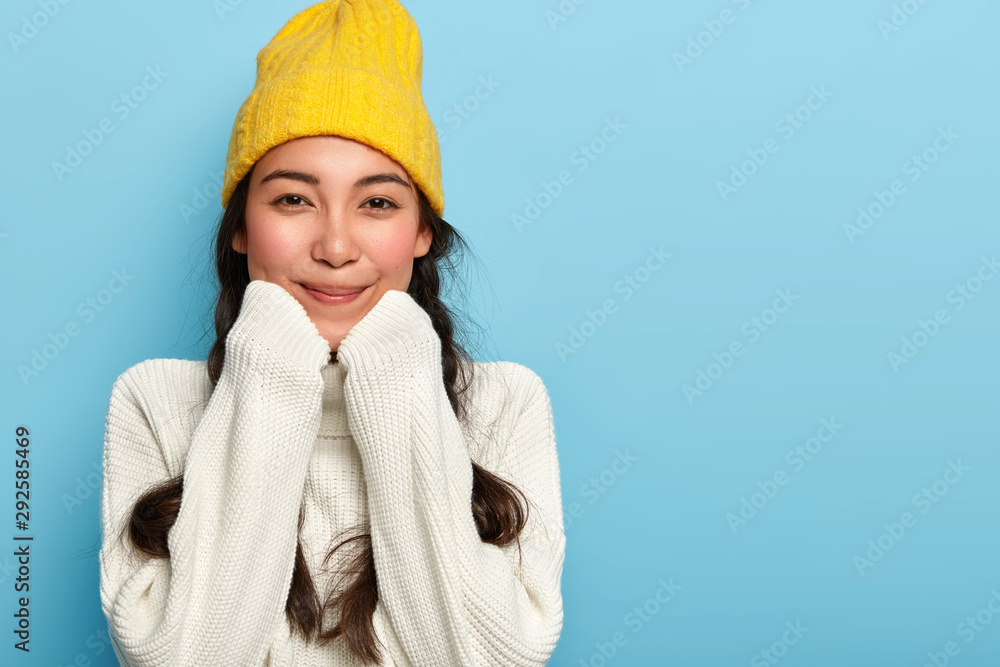 Portrait of delighted adorable Asian woman keeps hands under chin, dressed in oversized white sweater and yellow hat, enjoys comfort and calm domestic atmosphere, stays at home during cold weather - obrazy, fototapety, plakaty 