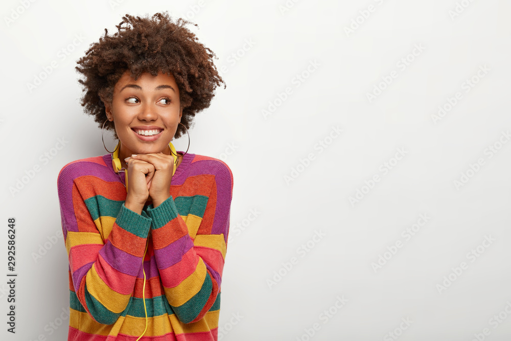 Horizontal shot of pleasant looking African American woman keeps hands under chin, looks away, dressed in colorful striped jumper, uses headphones for listening favourite music. Lifestyle concept Horizontal shot of pleasant looking African American woman keeps hands under chin, looks away, dressed in colorful striped jumper, uses headphones for listening favourite music. Lifestyle concept