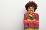 Portrait of pleased millennial Afro girl enjoys spare time, listens music via headphones, looks happily at you, gets pleasant news, wears striped sweater, poses against white studio wall, blank space